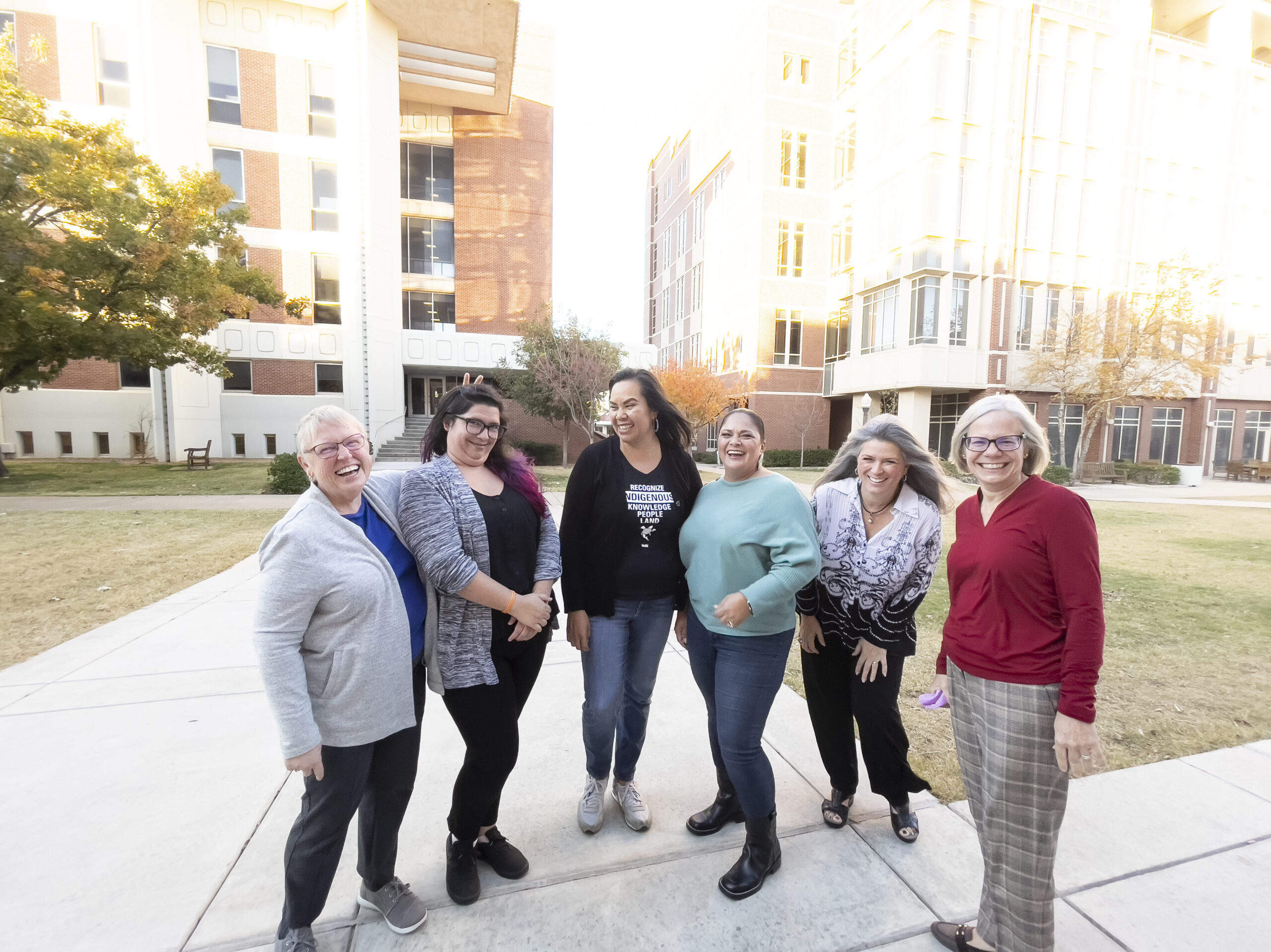 Picture of six faculty members smiling and laughing in front of Carson Engineering on the OU campus. Devon Energy Hall is in the background.